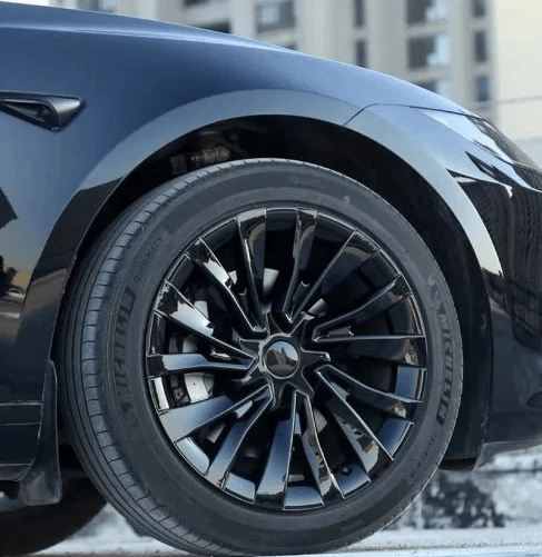 Close-up of a black car wheel with a glossy multi-spoke alloy rim and Michelin tires, parked outdoors with a blurred building in the background. TESLA MODEL 3 HIGHLAND 18 INCH WHEEL COVERS UBER NOVA PERFORMANCE STYLE GLOSS BLACK FREE DELIVERY WITH FEDEX ACROSS EUROPE FROM IRELAND AND EUROPES TESLARY.EU THE TESLA ACCESSORIES SPECIALISTS